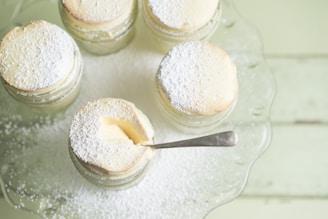 A collection of small souffle desserts in glass jars, topped with powdered sugar. One jar has a spoon inserted, indicating that it is ready to be eaten. The desserts are arranged on a glass tray placed on a light green wooden surface.