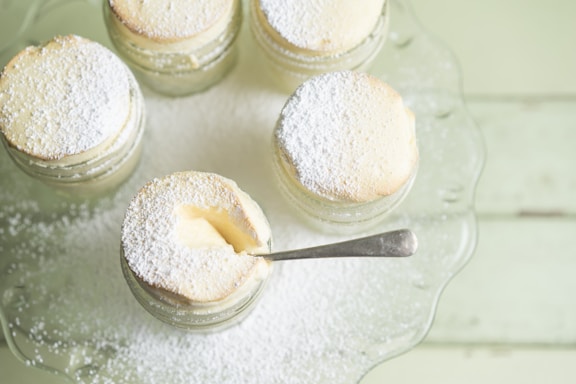 A collection of small souffle desserts in glass jars, topped with powdered sugar. One jar has a spoon inserted, indicating that it is ready to be eaten. The desserts are arranged on a glass tray placed on a light green wooden surface.