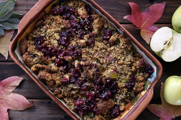 An appetizing baked dish with a crumbly, golden-brown topping and scattered dark red cranberries. The dish is presented in a large, rectangular earthenware baking dish. Surrounding the dish are vibrant autumn leaves and freshly sliced and whole green apples on a wooden surface.