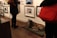 A gallery setting with framed photographs and artworks displayed on the wall. A brindle-colored dog is lying on a green mat on the wooden floor. People are standing around, wearing coats and holding bags, observing the art.