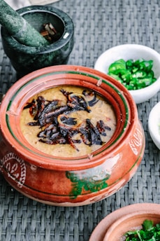 A clay pot filled with a creamy dish topped with caramelized onions or spices is placed on a woven surface. Surrounding the pot is a stone mortar containing some spices, a small white bowl with sliced green chili peppers, and another clay pot partially visible.
