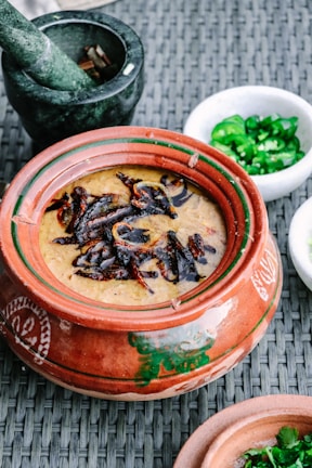 A clay pot filled with a creamy dish topped with caramelized onions or spices is placed on a woven surface. Surrounding the pot is a stone mortar containing some spices, a small white bowl with sliced green chili peppers, and another clay pot partially visible.