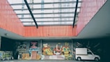 An urban setting featuring vibrant graffiti art on a wall under a large glass-covered atrium. The artwork includes colorful characters and abstract designs. A white truck is parked to the right side, and the surrounding architecture includes tall buildings in the background.