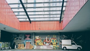 An urban setting featuring vibrant graffiti art on a wall under a large glass-covered atrium. The artwork includes colorful characters and abstract designs. A white truck is parked to the right side, and the surrounding architecture includes tall buildings in the background.