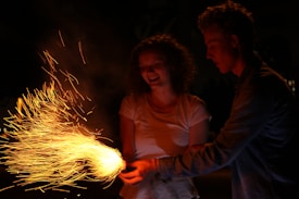 A young woman and a young man are together in the dark, holding a sparkler that emits bright, fiery sparks in the air. The woman is smiling brightly, suggesting a joyful atmosphere.