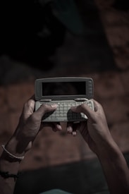 Two hands holding an older model mobile phone with a full QWERTY keyboard. The phone is open revealing the keyboard and a small display screen. The lighting is dim, giving a nostalgic and somewhat somber atmosphere.
