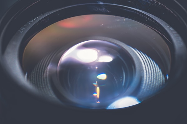 A close-up view of a camera lens, focusing on the intricate glass elements that reflect and refract light. The lens surface shows various reflections and highlights.