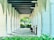 An outdoor corridor with white stone arches creates a perspective leading to a bright opening filled with greenery. A young child, dressed in a light-colored outfit, stands beside a stroller, possibly examining something on a wall. Overhead, black industrial-style lamps are symmetrically aligned, contributing to the elegance of the architectural design.