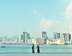 A clear view of a city skyline across a calm body of water, featuring modern skyscrapers with visible logos, under a lightly clouded sky. Two individuals stand on a waterfront promenade, gazing out towards the city.