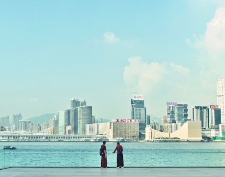 A clear view of a city skyline across a calm body of water, featuring modern skyscrapers with visible logos, under a lightly clouded sky. Two individuals stand on a waterfront promenade, gazing out towards the city.