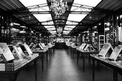 A spacious indoor market or bookstore with rows of tables displaying stacks of books. The ceiling features a metal framework with chandeliers hanging down, providing a sense of elegance. The setting has a structured, symmetrical layout with books standing upright and arranged uniformly on each table.