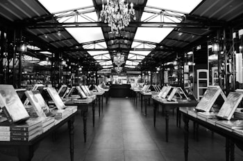 A spacious indoor market or bookstore with rows of tables displaying stacks of books. The ceiling features a metal framework with chandeliers hanging down, providing a sense of elegance. The setting has a structured, symmetrical layout with books standing upright and arranged uniformly on each table.