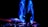 A dynamic and colorful light display is happening with vibrant blue trails swirling against a dark background. Silhouettes of people and tables topped with candles are visible under large umbrellas, indicating a possible event or gathering.