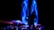 A dynamic and colorful light display is happening with vibrant blue trails swirling against a dark background. Silhouettes of people and tables topped with candles are visible under large umbrellas, indicating a possible event or gathering.