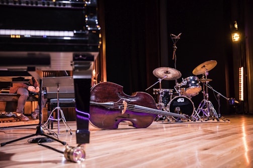 A stage setup is visible with several musical instruments including a grand piano, a double bass lying on its side, and a drum set. To the left, a person is seated with legs crossed, holding what appears to be a laptop or a book. The stage floor is wooden, and the background is dark with some stage lighting visible.