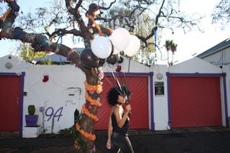 A person with a large afro hairstyle is holding a handful of black and white balloons. They are standing in front of a vividly decorated tree with colorful ribbons. The background features a white wall with purple trim, red garage doors, and house number 94 written on it. It's a sunny day with clear skies.