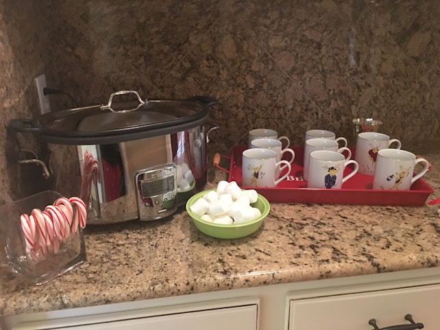 A kitchen countertop with a large slow cooker, a container of candy canes, a small green bowl of marshmallows, and a red tray holding nine white mugs with whimsical designs. The countertop is made of granite with a speckled pattern.