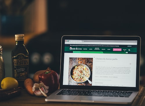 An Apple MacBook Pro with a recipe from the Jamie Oliver website on a kitchen counter, with veg and Filippo Berio olive oil n