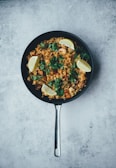 A skillet filled with a vibrant dish consisting of rice, garnished with fresh cilantro and sliced lemon wedges. The rice is mixed with various colorful ingredients, possibly including shrimp or chicken, and set against a textured gray background.