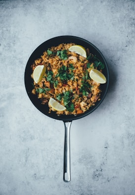 A skillet filled with a vibrant dish consisting of rice, garnished with fresh cilantro and sliced lemon wedges. The rice is mixed with various colorful ingredients, possibly including shrimp or chicken, and set against a textured gray background.