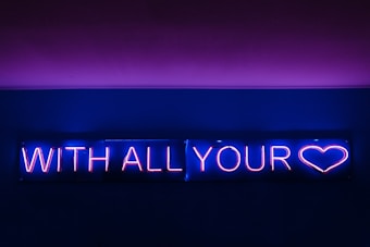 A neon sign glowing in bright pink and blue colors displays the phrase 'WITH ALL YOUR' followed by the outline of a heart.