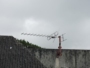 A rooftop antenna with a rusted metal base is mounted on a textured concrete wall. The sky is overcast, contributing to a muted, gloomy ambiance. In the background, a line of trees is partially visible.