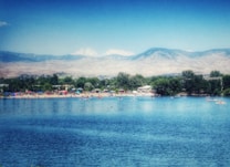 A serene lake with vibrant blue water reflecting a clear sky. In the distance, people enjoy a sunny day at a sandy beach dotted with colorful umbrellas and tents. Verdant trees line the shore, with mountains rising majestically in the background under a light haze.
