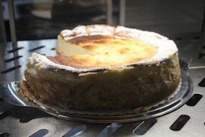 A large, round cheesecake with a golden-brown top and a thick crust edges sits on a metal tray. The top has a slightly cracked surface, dusted with powdered sugar.