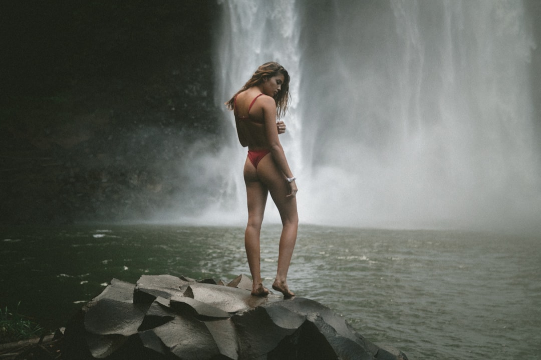 woman in red panty and bra standing of black stone near waterfalls