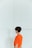 A person with short dark hair is standing against a white wall, wearing a bright orange shirt and denim jeans. The person is turned slightly away, revealing large hoop earrings and part of a printed design on the shirt.