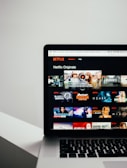 A laptop displaying the Netflix Originals section with various show thumbnails, including Stranger Things, Black Mirror, and Riverdale. The screen interface includes a list of shows available for viewing.