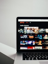 A laptop displaying the Netflix Originals section with various show thumbnails, including Stranger Things, Black Mirror, and Riverdale. The screen interface includes a list of shows available for viewing.
