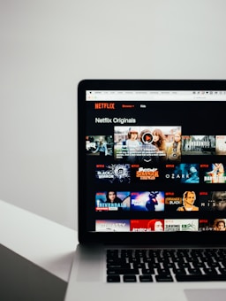 A laptop displaying the Netflix Originals section with various show thumbnails, including Stranger Things, Black Mirror, and Riverdale. The screen interface includes a list of shows available for viewing.