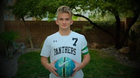 A person wearing a white sports jersey with the number 7 and the word Panthers printed on it. They are holding a blue and green soccer ball in both hands. In the background, there is a wooden fence and some trees, suggesting an outdoor setting.