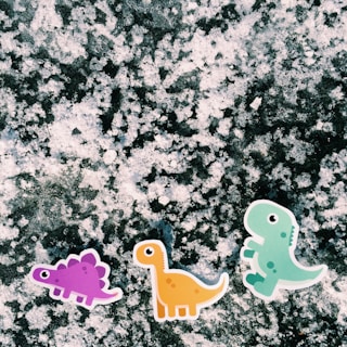 Three cartoon dinosaur stickers, each a different color, are placed on a snowy surface. The background is textured with a mottled appearance, giving it a frosty or icy look.
