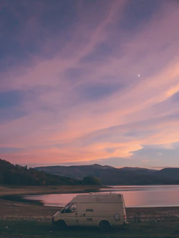 A serene motorhome parked beside a calm lake at sunset, surrounded by lush greenery.