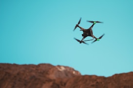 A drone with four rotors is flying against a clear blue sky above a rocky landscape. The drone has bright green lights on its front arms.