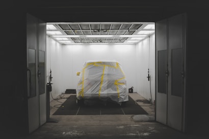 A vehicle is positioned in a spacious, enclosed area resembling a spray booth. It is covered with white material secured by yellow tape, suggesting preparation for painting. The environment is lit by bright overhead lights, and equipment like hoses and a spray gun are visible, indicating an automotive workshop setting.