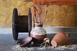 An arrangement of rustic items placed indoors against a textured wooden wall. A large spool rests upright on the left, displaying some greenery. A brown pottery vase with white zigzag patterns is filled with decorative dried stems. Beside it lies a shallow wooden bowl and a larger clay pot on its side with scattered straw and a small bouquet of colorful flowers nearby on the concrete floor.