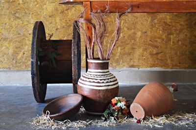 An arrangement of rustic items placed indoors against a textured wooden wall. A large spool rests upright on the left, displaying some greenery. A brown pottery vase with white zigzag patterns is filled with decorative dried stems. Beside it lies a shallow wooden bowl and a larger clay pot on its side with scattered straw and a small bouquet of colorful flowers nearby on the concrete floor.