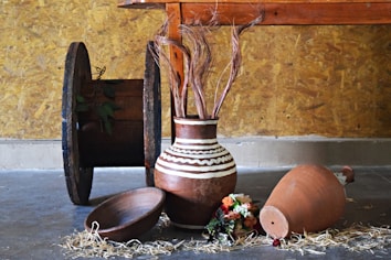 An arrangement of rustic items placed indoors against a textured wooden wall. A large spool rests upright on the left, displaying some greenery. A brown pottery vase with white zigzag patterns is filled with decorative dried stems. Beside it lies a shallow wooden bowl and a larger clay pot on its side with scattered straw and a small bouquet of colorful flowers nearby on the concrete floor.