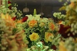 A lush and intricate floral arrangement featuring a variety of blooms including white lilies, orange and yellow roses, and sprigs of baby’s breath. The composition is vibrant and dense, set against a dimly lit background with hints of chandeliers above, creating a sophisticated ambiance.