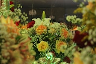 A lush and intricate floral arrangement featuring a variety of blooms including white lilies, orange and yellow roses, and sprigs of baby’s breath. The composition is vibrant and dense, set against a dimly lit background with hints of chandeliers above, creating a sophisticated ambiance.