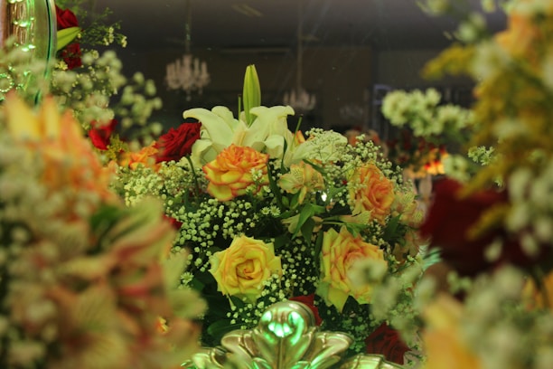A lush and intricate floral arrangement featuring a variety of blooms including white lilies, orange and yellow roses, and sprigs of baby’s breath. The composition is vibrant and dense, set against a dimly lit background with hints of chandeliers above, creating a sophisticated ambiance.