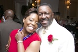 A smiling couple poses closely together in a formal setting. The woman is wearing a red dress with a blue necklace, while the man is dressed in a white suit with a red rose boutonniere. Other guests can be seen in the background, suggesting this is a social event.