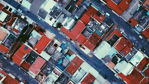 An aerial view of a densely packed urban neighborhood with a grid-like pattern of streets and buildings. Red roofs dominate the scene, interspersed with gray and blue rooftops. Cars are visible on the streets, which run between rows of structures. The perspective highlights the orderly arrangement and repetition of shapes.