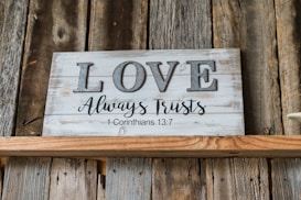 A rustic wooden sign is displayed against a background of weathered wood planks. The sign features the word 'LOVE' in large, gray, metallic letters, followed by the phrase 'Always Trusts' and '1 Corinthians 13:7' in elegant script.