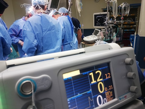 A group of medical professionals wearing blue surgical attire performing a surgery. In the foreground, there is a medical monitor displaying vital signs. The background includes medical equipment like IV drips, monitors, and respiratory devices. The setting appears to be a hospital operating room.