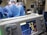 A group of medical professionals wearing blue surgical attire performing a surgery. In the foreground, there is a medical monitor displaying vital signs. The background includes medical equipment like IV drips, monitors, and respiratory devices. The setting appears to be a hospital operating room.