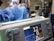 A group of medical professionals wearing blue surgical attire performing a surgery. In the foreground, there is a medical monitor displaying vital signs. The background includes medical equipment like IV drips, monitors, and respiratory devices. The setting appears to be a hospital operating room.